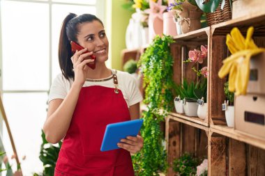 Young beautiful hispanic woman florist talking on smartphone using touchpad at flower shop