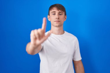 Kafkasyalı sarışın adam mavi arka planda duruyor. Parmağını kaldırmış ve kızgın bir ifadeyle işaret ediyor. 