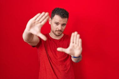 Young hispanic man standing over red background doing frame using hands palms and fingers, camera perspective 