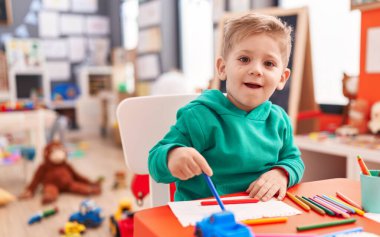 Sevimli beyaz çocuk anaokulunda masa başında oturmuş not defterine resim çiziyor.