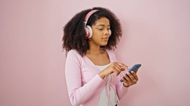 Akıllı telefon ve kulaklık kullanan Afro-Amerikalı kadın izole edilmiş pembe arka plan üzerinde ciddi bir ifade kullanıyor.