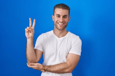 Mavi arka planda dikilmiş, gülümseyen ve kameraya göz kırpan beyaz bir adam parmaklarıyla zafer işareti yapıyor. İki numara.. 