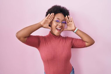 Beautiful african woman with curly hair standing over pink background smiling cheerful playing peek a boo with hands showing face. surprised and exited 