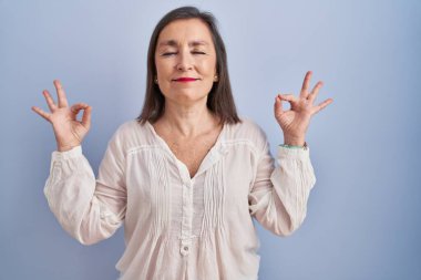 Orta yaşlı Latin bir kadın mavi arka planda durup rahatlamış ve parmaklarıyla meditasyon hareketi yaparken gözleri kapalı gülümsüyor. yoga konsepti. 