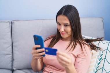 Akıllı telefon ve kredi kartı kullanan genç, güzel İspanyol kadın evdeki koltukta oturuyor.