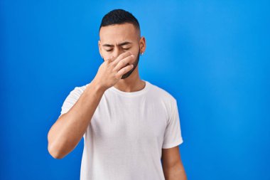 Young hispanic man standing over blue background smelling something stinky and disgusting, intolerable smell, holding breath with fingers on nose. bad smell 