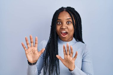 Afro-Amerikalı bir kadın mavi arka planda korku ve korku içinde duruyor. El hareketlerini durdur, şok içinde bağır. panik konsepti. 