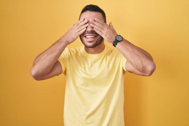 Sarı arka planda dikilen genç İspanyol adam, neşeli ve komik gülümseyen ellerle gözlerini kapatıyor. kör kavram. 