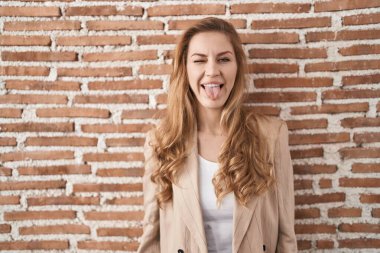 Güzel sarışın kadın tuğla duvarın üzerinde duruyor. Dilini komik bir ifadeyle dışarı çıkarıyor. duygu kavramı. 