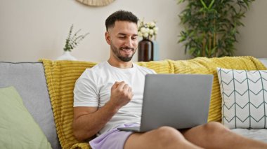 Genç Arap adam dizüstü bilgisayar kullanıyor. Kanepede oturmuş kutlama yapıyor.