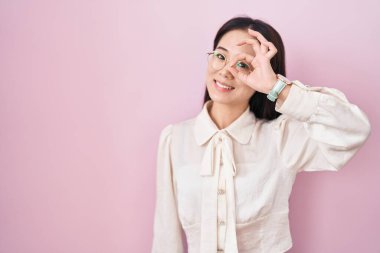 Pembe arka planda duran genç Çinli kadın el hareketleriyle gülümsüyor ve mutlu yüzüyle parmaklarının arasından bakıyor.. 