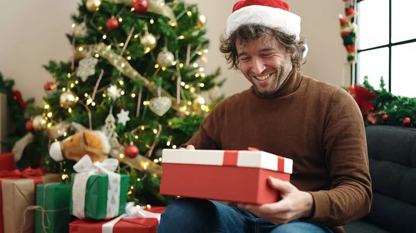 自宅でクリスマスツリーでソファーに座っている贈り物を見ている若いヒスパニックの男 — ストック写真