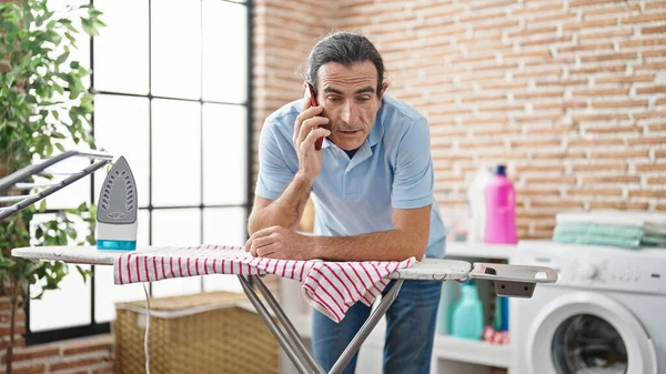 Middle age man talking on smartphone leaning on ironing machine at laundry room