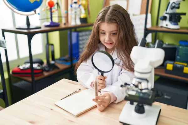 Adorable hispanic girl student looking sample with magnifying glass at laboratory classroom