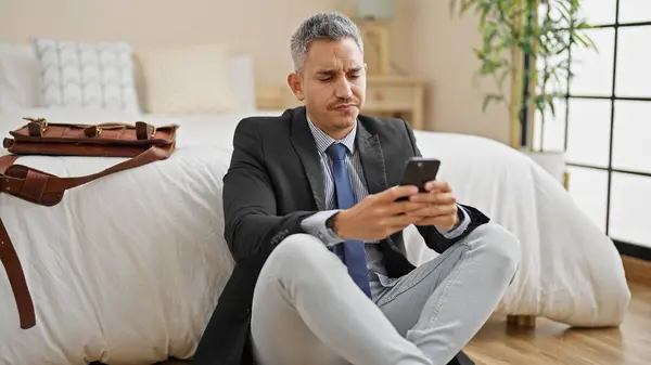 Young hispanic man business worker using smartphone sitting on floor at hotel room