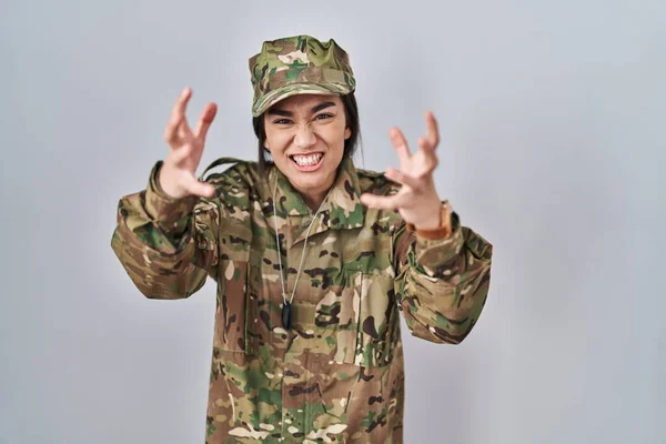 Young south asian woman wearing camouflage army uniform shouting frustrated with rage, hands trying to strangle, yelling mad