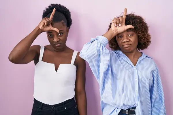 ピンクの背景の上に立っている2人のアフリカの女性は 負け犬のジェスチャーを嘲笑し侮辱するために額に指を持つ人々の楽しみを作ります — ストック写真