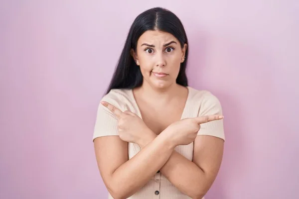 年轻的惊慌失措的女人站在粉色的背景上 用手指指指两边 方向不一 — 图库照片