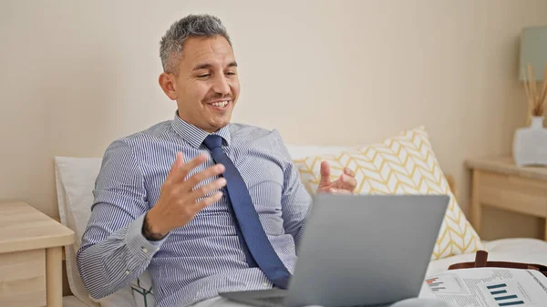 Young hispanic man business worker having video call sitting on bed at hotel room