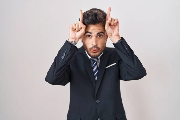 ビジネスススーツを着てタトゥーを着た若いヒスパニック男性とブルホーンとして頭の上に指で面白いジェスチャーをするタイ — ストック写真