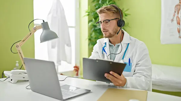 Young caucasian man doctor on video call writing on clipboard at clinicc
