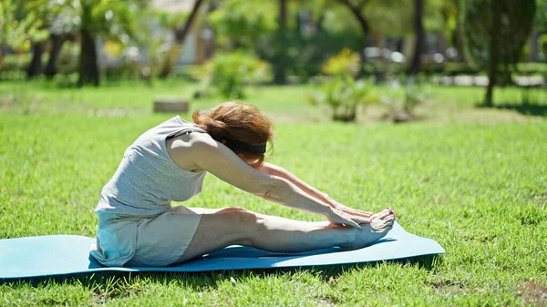 Middle age woman stretching legs sitting on yoga mat at park