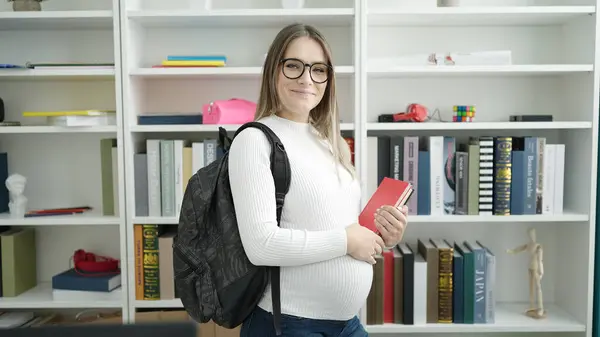在图书馆大学 年轻的怀孕女生带着自信的笑容举着书本 — 图库照片