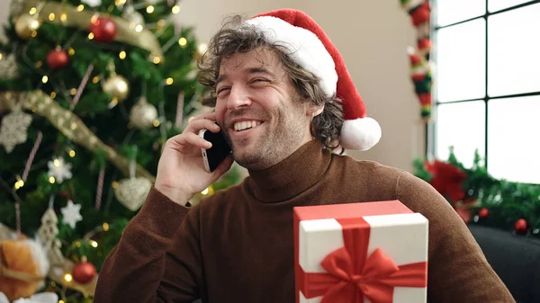 若いヒスパニックの男は 自宅でクリスマスプレゼントを持ってソファーに座ってスマートフォンで話します — ストック写真