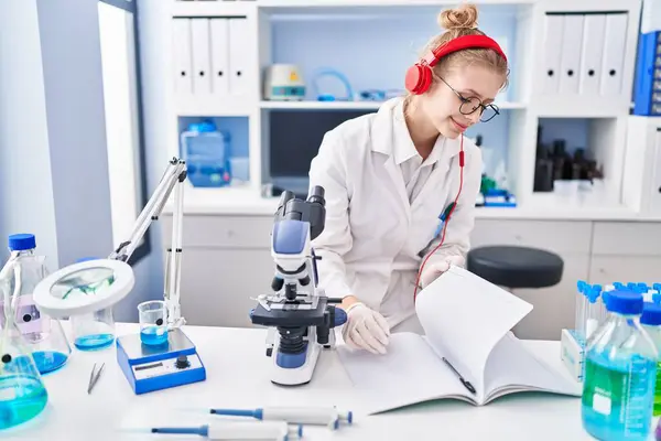 Young blonde woman scientist listening to music writing on notebook at laboratory