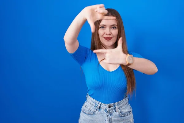 青い背景の上に立っている赤毛の女性はハッピーフェイスと手と指でフレームを作る笑顔 クリエイティビティと写真コンセプト — ストック写真