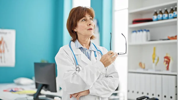 Middle age woman doctor standing with serious expression and arms crossed gesture holding glasses at clinic