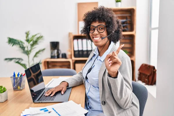 Black woman with curly hair wearing call center agent headset at the office with a big smile on face, pointing with hand finger to the side looking at the camera.