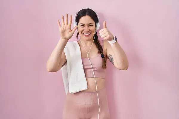 Young brunette woman wearing sportswear and headphones showing and pointing up with fingers number six while smiling confident and happy.