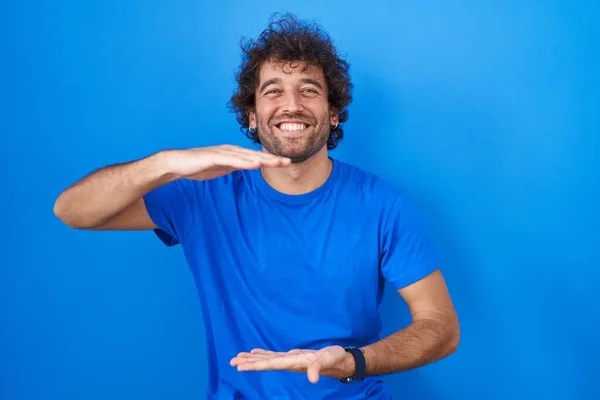 西班牙裔年轻人站在蓝色背景的旁边 手举着大号和大号的手势 测量符号 微笑着看着摄像机 计量概念 — 图库照片