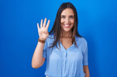 Genç esmer kadın mavi arka planda duruyor ve parmaklarını dört numarayı gösterip işaret ederken kendinden emin ve mutlu bir şekilde gülümsüyor.. 