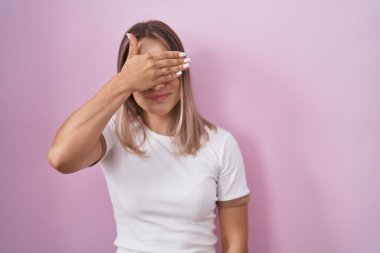 Sarışın, beyaz bir kadın pembe arka planda duruyor. Gülümsüyor ve elleri yüzünde, şaşırtmak için gözleri örtüyor. kör kavram. 