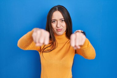 Young brunette woman standing over blue background punching fist to fight, aggressive and angry attack, threat and violence 