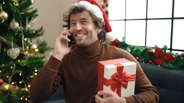 Genç İspanyol adam akıllı telefonuyla konuşuyor. Kanepede oturuyor. Elinde Noel hediyesi var.