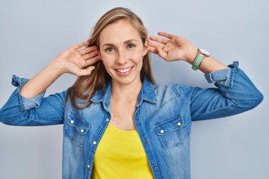 Mavi arka planda duran genç sarışın kadın gülümsüyor parmaklarıyla kulaklarını çekiyor, komik bir jest. deneme sorunu 