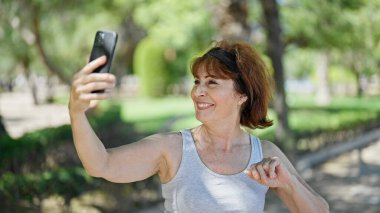 Orta yaşlı bir kadın, parktaki akıllı telefondan özçekim yaparken kendinden emin bir şekilde gülümsüyor.