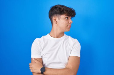 Mavi arka planda duran İspanyol genç kollarını kavuşturmuş ve kendinden emin bir şekilde yan tarafa bakıyor. 