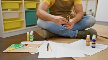 Genç Latin ressam sanat stüdyosunda elinde fırçalarla yerde oturuyor.