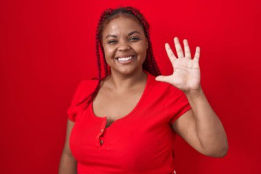 Kırmızı arka planda örülmüş saçları olan Afro-Amerikalı bir kadın beş numaralı parmaklarıyla gülümseyerek kendinden emin ve mutlu bir şekilde duruyor.. 