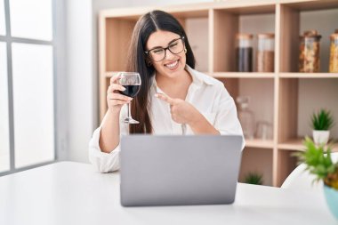 Genç esmer kadın dizüstü bilgisayarla video görüşmesi yapıyor. Bir bardak şarap içiyor. Gülümsüyor. 