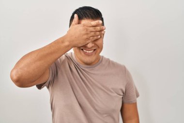 Latin kökenli genç bir adam beyaz arka planda duruyor. Gülümsüyor ve gülümsüyor. Gözleri şaşkınlıkla örtüyor. kör kavram. 