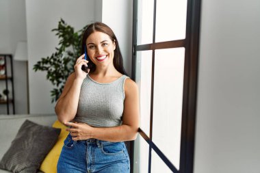 Genç, güzel, İspanyol bir kadın akıllı telefondan konuşuyor. Evde duruyor.