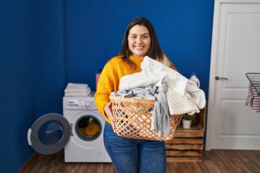 Young beautiful plus size woman smiling confident holding basket with clothes at laundry room