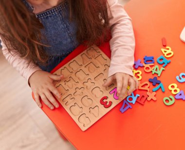 Adorable hispanic girl playing with maths puzzle game sitting on table at kindergarten