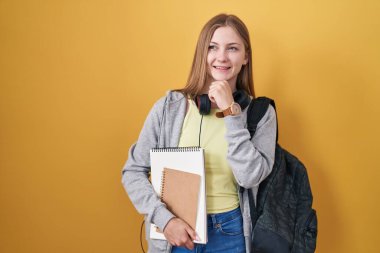 Genç beyaz bir kadın öğrenci çantası takıyor ve elleri çenesinde kitapları tutuyor. Soru sormayı düşünüyor. Düşünceli bir ifade. Gülümseyen ve düşünceli bir yüz. şüphe kavramı. 