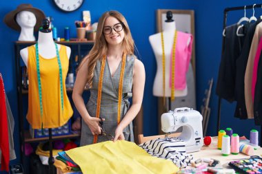 Young woman tailor smiling confident cutting cloth at clothing factory clipart